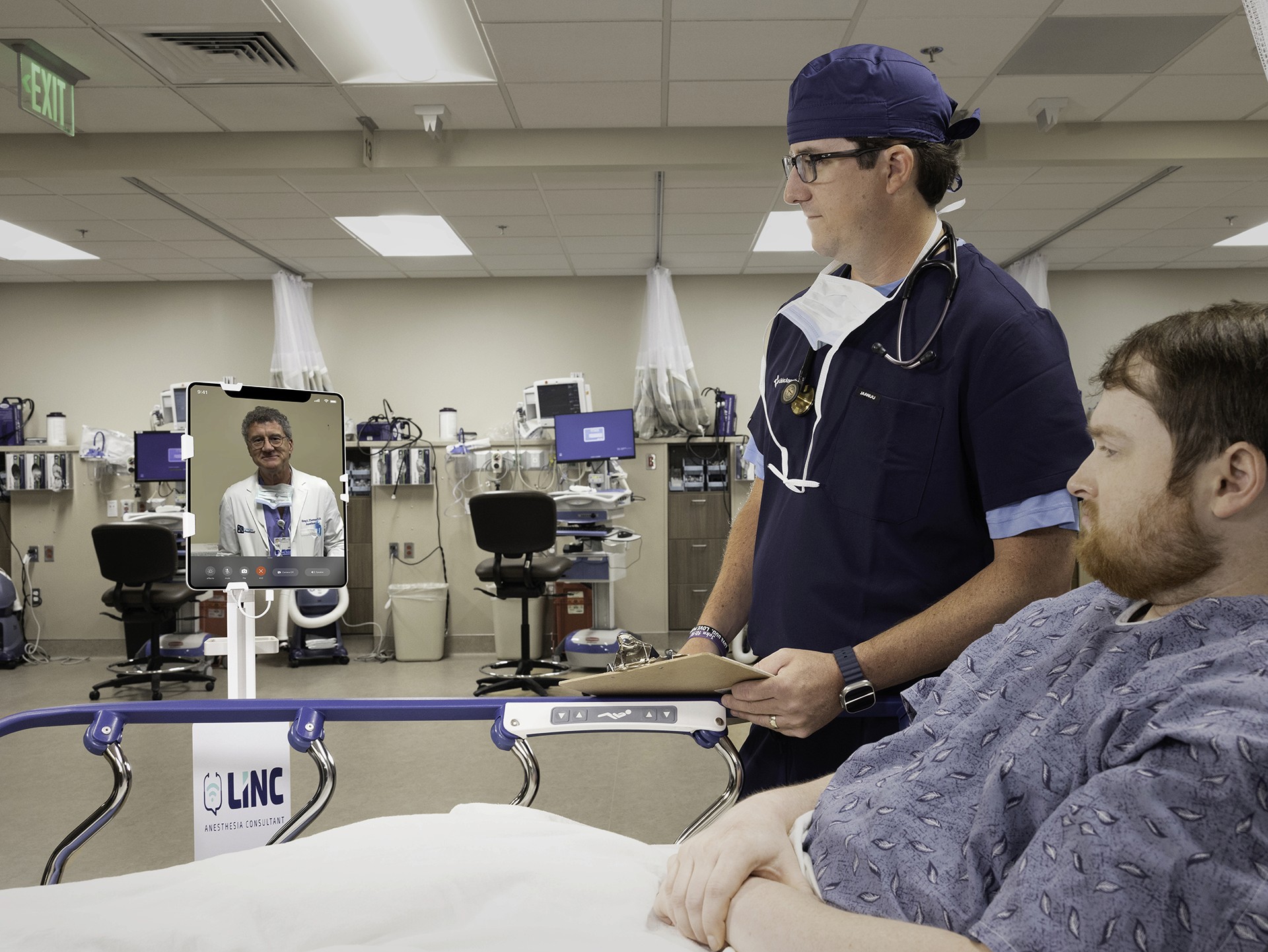 A CRNA in scrubs uses the LINC teleconsultation tablet at the bedside in a clinical setting, representing virtual access to experienced anesthesiologists for real-time support