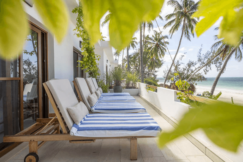 Row of wooden sun loungers with blue-and-white striped towels on a white balcony overlooking a tropical beach with palm trees and ocean views.