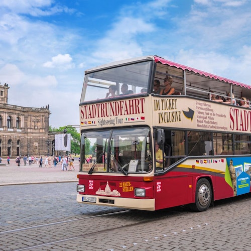 Kaksikerroksinen sightseeing-bussi, jonka kyljessä on saksankielinen teksti, historiallisella kaupunkiaukiolla, jossa on ihmisiä ja taustalla koristeellinen rakennus.