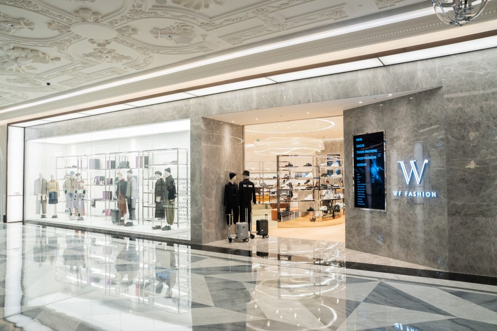 Exterior view of a modern fashion store labeled 'WF FASHION' featuring a sleek glass display of clothing and accessories, with polished marble flooring and elegant interior design.
