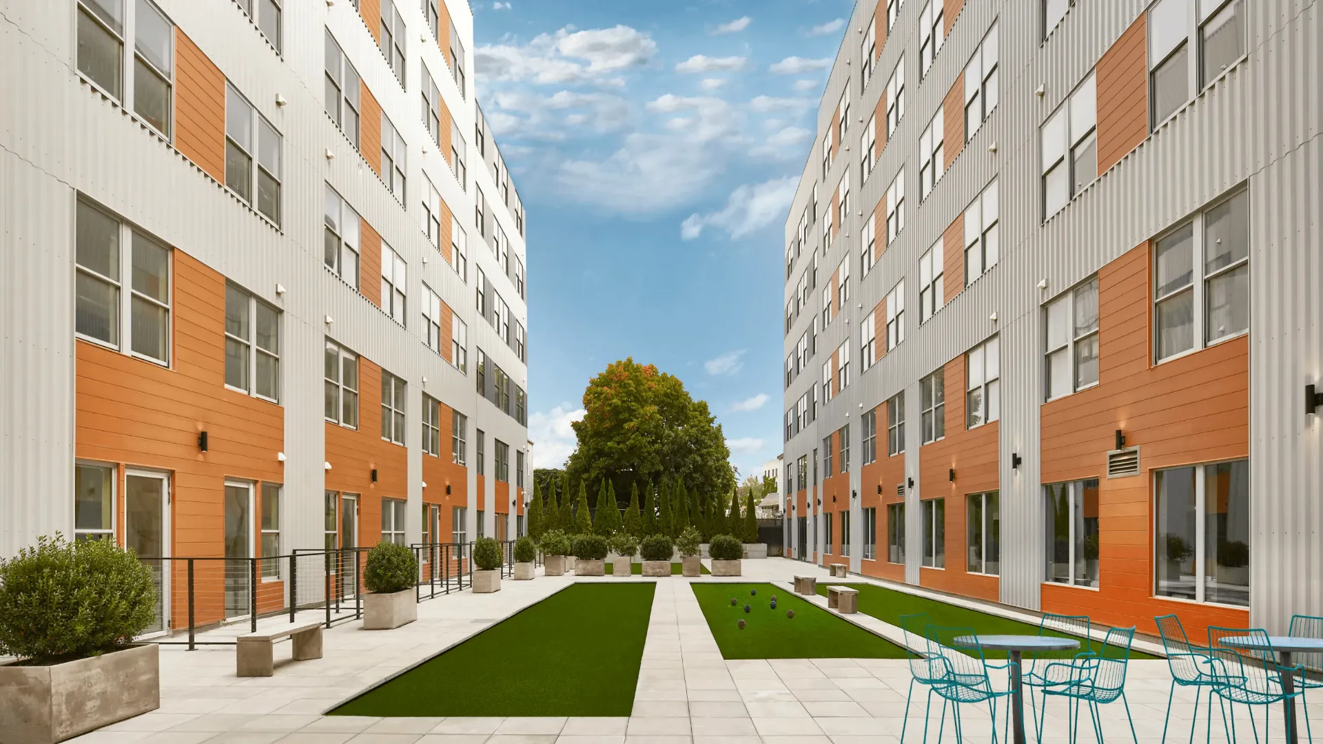 A modern courtyard view between orange-accented buildings under a clear blue sky, featuring green grass and trees.