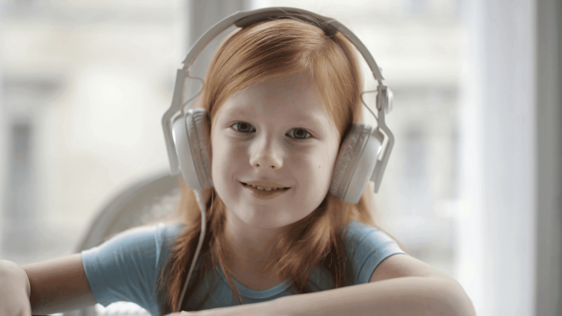 A young girl sitting on a grey blanket, wearing headphones and holding a phone, smiling while listening to HushAway®’s Sound Sanctuary.