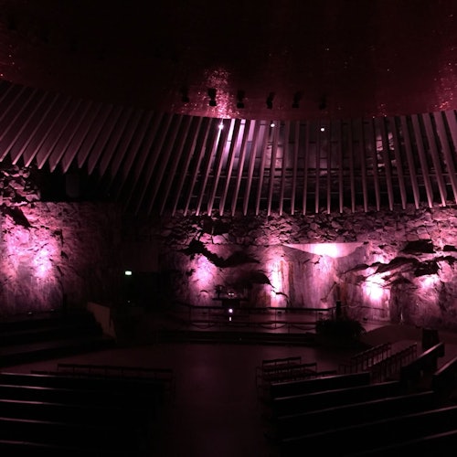 Hämärästi valaistu auditorio, jossa on kiviseinät, violetit valot ja porrastetut istuimet. Suuret kylkiluun muotoiset palkit kaartuvat lavan yläpuolelle.