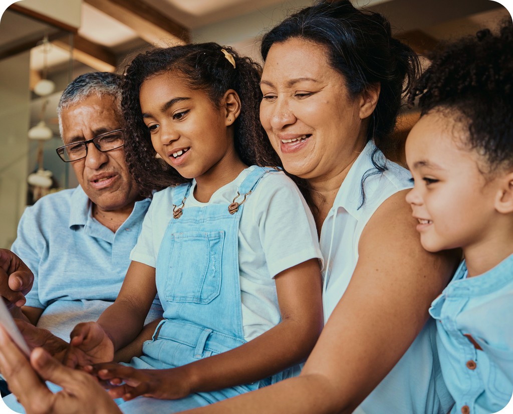 A smiling, multigenerational family group—a grandfather, grandmother, and two granddaughters—looking at a mobile device together