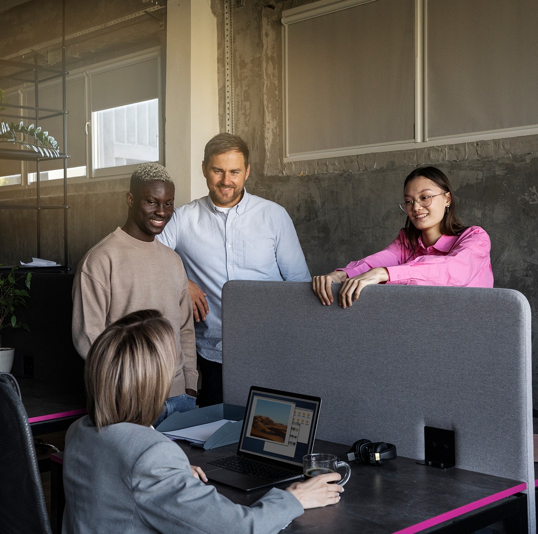 A diverse group of four people in a modern office setting gather around a desk where one person sits with an open laptop displaying a landscape image.