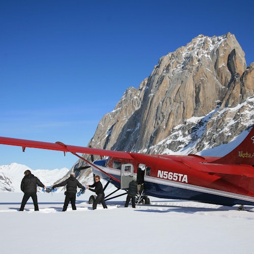 四個人站在一架紅色的有滑雪板的飛機旁，背景是被雪覆蓋的地面和白雪皚皚的山脈。