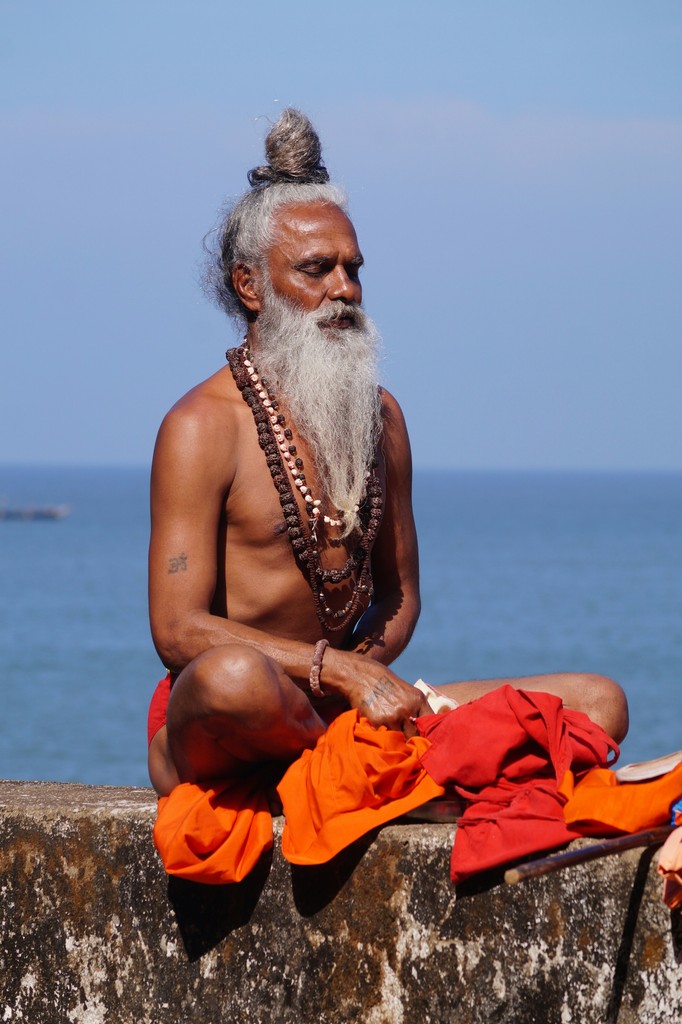 An elderly Indian sage with a long white beard and tied-up hair sits in meditation by the sea, adorned with beaded necklaces and orange robes, embodying spirituality and peaceful reflection.