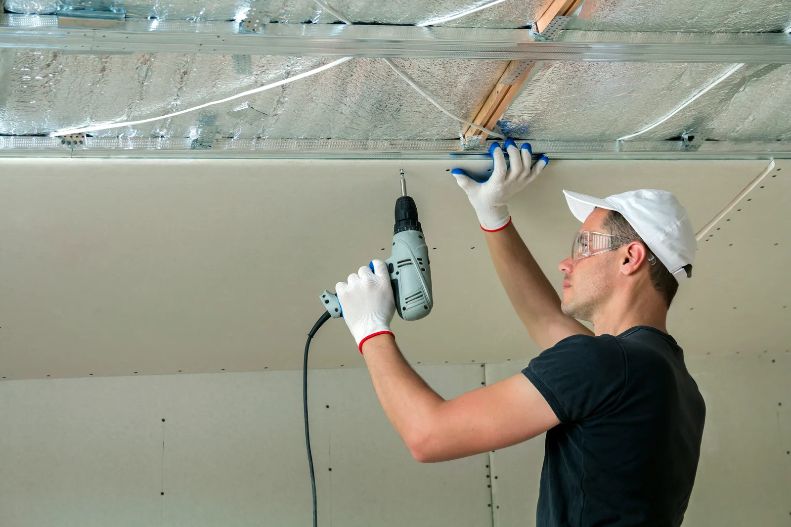 Worker drilling drywall ceiling panel onto insulated metal frame