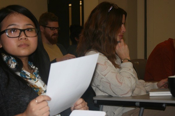A photo of a woman wearing glasses while siting in a classroom and holding a piece of paper.