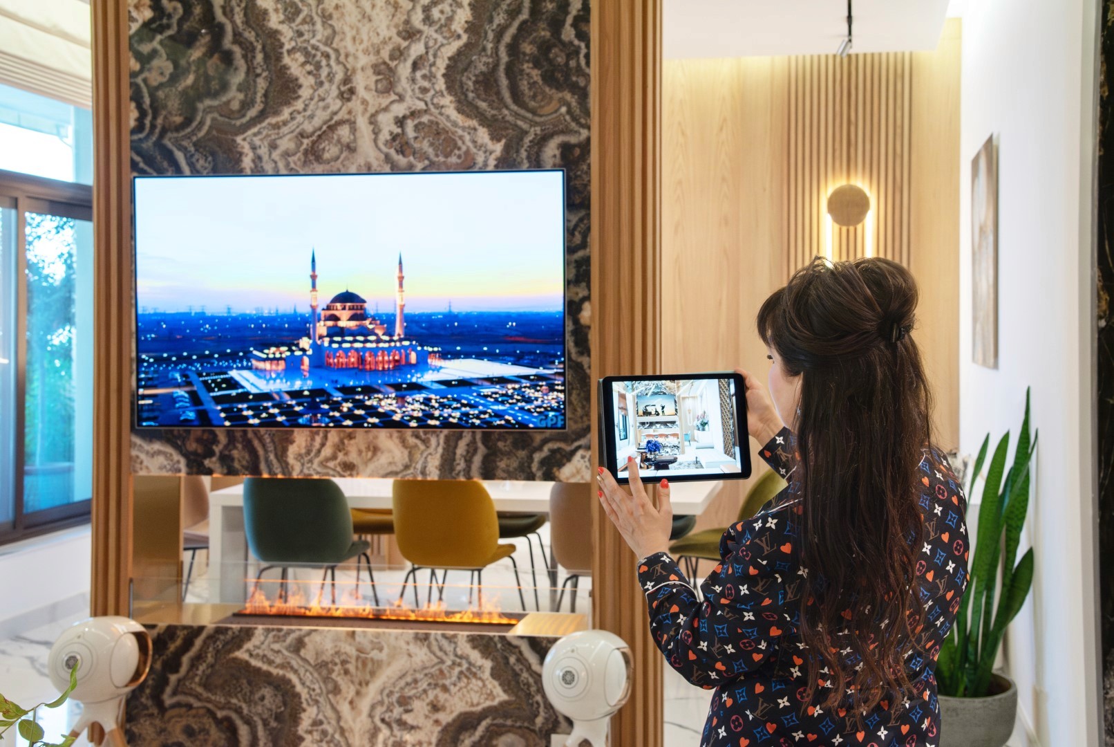 Mrs Lora Bergiy showing renders stands by a marble-framed television displaying a vibrant image of a mosque at sunset, with a modern dining area in the background.