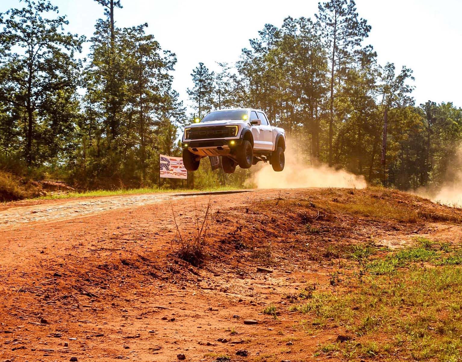 Ford F-150 Raptor jumping over a dirt trail, kicking up dust in a wooded off-road setting.