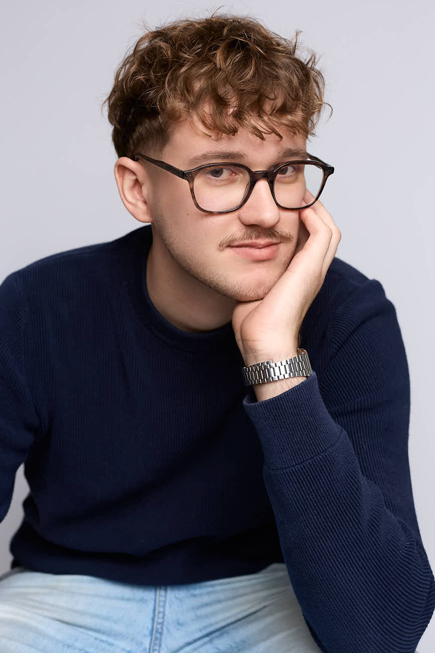 Portrait corporate en studio d'un designer graphique basé à Lille, portant des lunettes et un pull bleu marine, sur fond gris neutre.