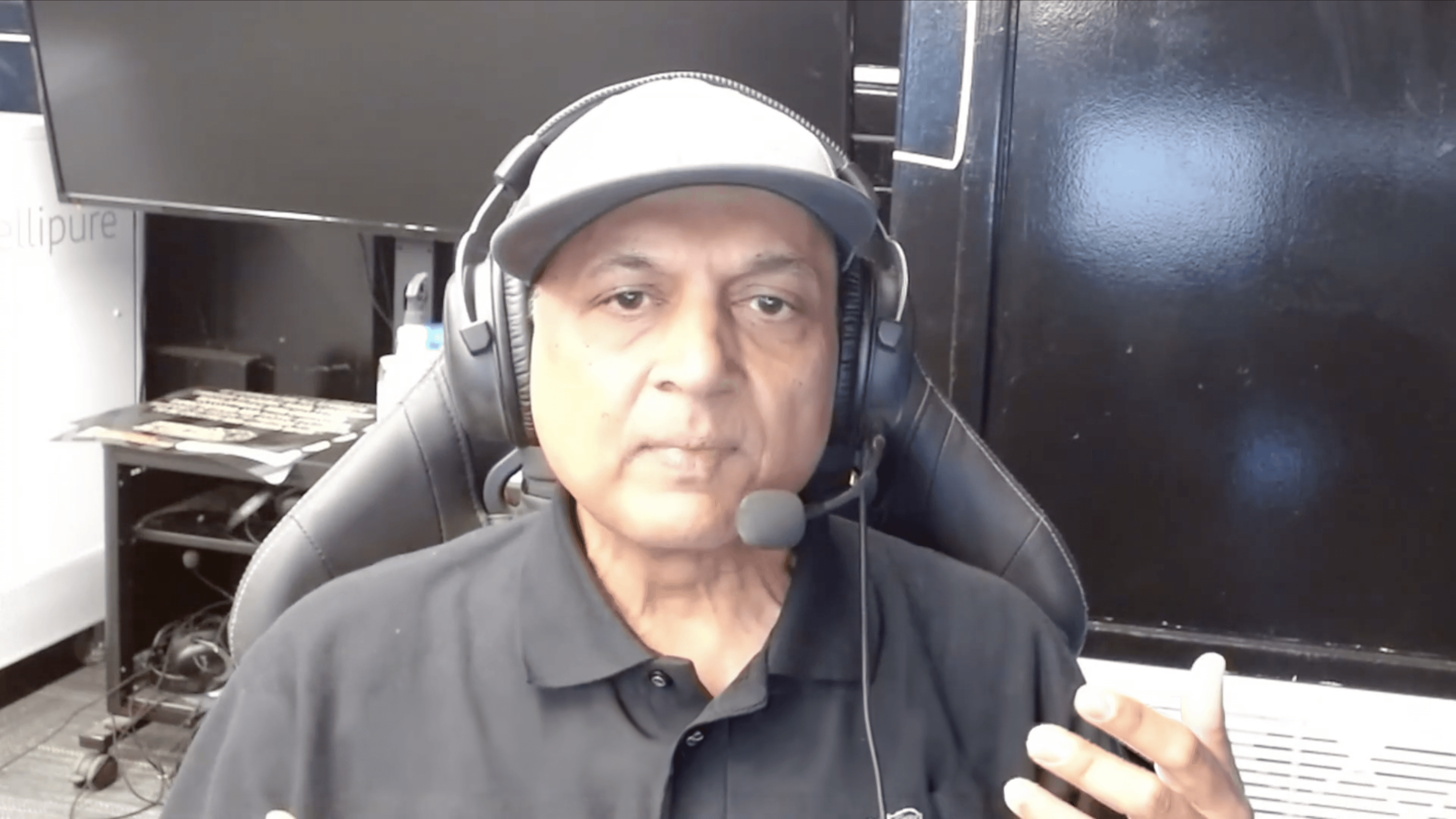 A man wearing a black polo shirt and a light-colored baseball cap sits in a high-backed chair, facing the camera. He is wearing over-ear headphones with an attached microphone, suggesting he is speaking or participating in an online meeting or recording. The background includes a dark cabinet and a desk with computer equipment, indicating a home office or studio setting.