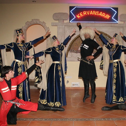 Artister i traditionella dräkter poserar under en neonskylt med texten "Kervansaray." Tre män, fyra kvinnor och en valvbåge är synliga.