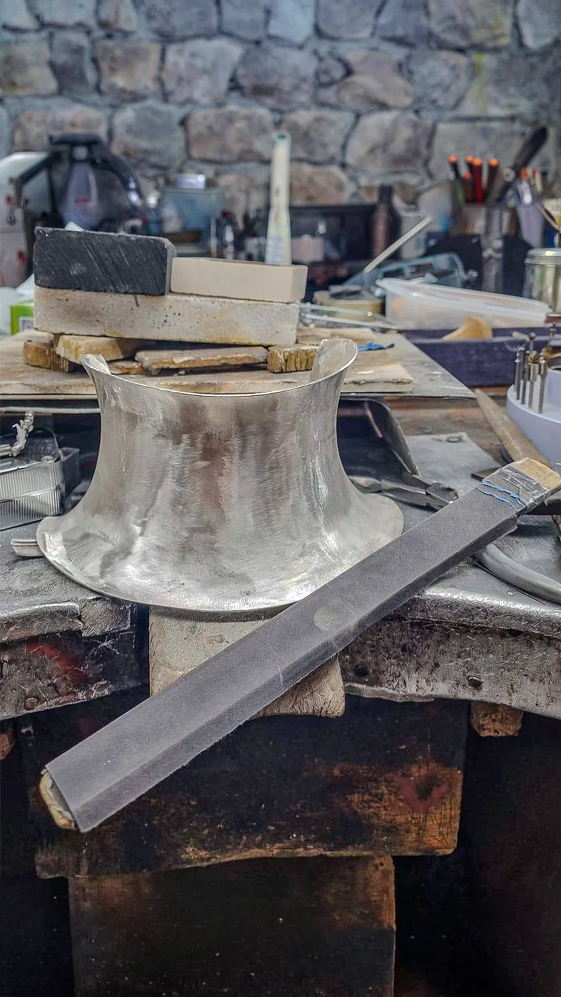 A high silver collar piece mid-craft sitting on a jeweler's workshop bench surrounded by artisan tools, files, and craft blocks, labeled "03 Form."