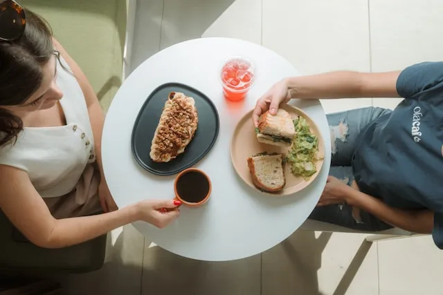 Top-down view of two people sharing a meal at a small round table with sandwiches and drinks.