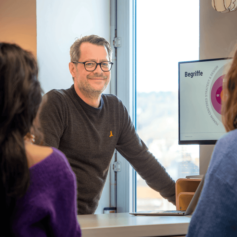 Mann mit Brille und dunkelgrauem Pullover lehnt an einem Tisch vor einem Bildschirm mit der Überschrift 'Begriffe' und mehreren Personen im Vordergrund.