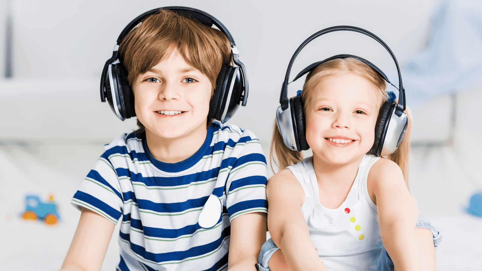 Two children sitting down, both wearing headphones and smiling at the camera while listening to HushAway®’s Sound Sanctuary.