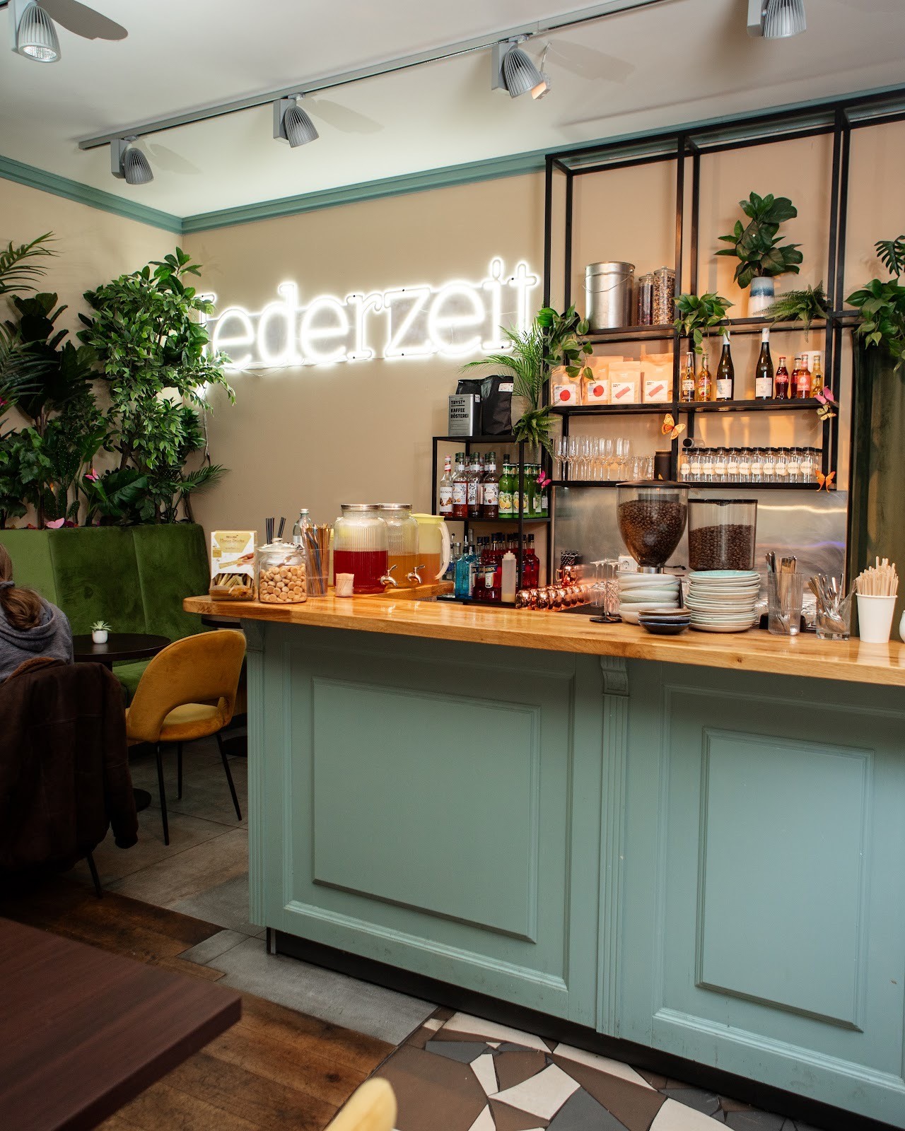 Interior view of Jederzeit Cafe showing a long, light blue counter with a barista working behind it, shelves with plants and products, and warm lighting.
