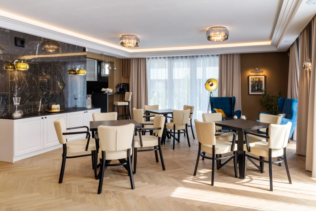 Elegancka jadalnia z kwadratowymi stołami i kremowymi krzesłami, w tle kuchnia z blatem z czarnego marmuru.
