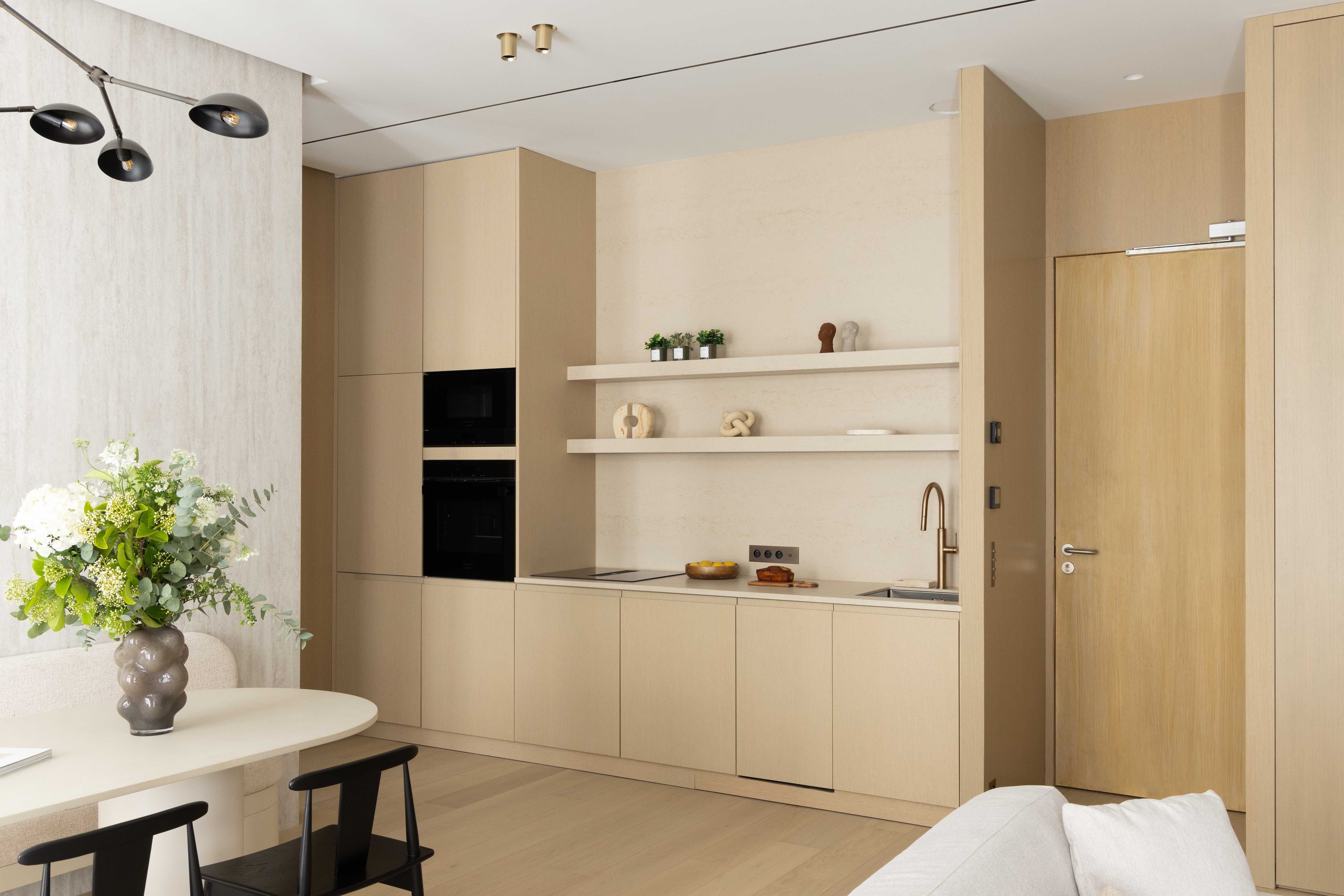 Cuisine ouverte aux facades beiges en bois clair avec four encastre noir, etageres blanches et table ronde avec bouquet