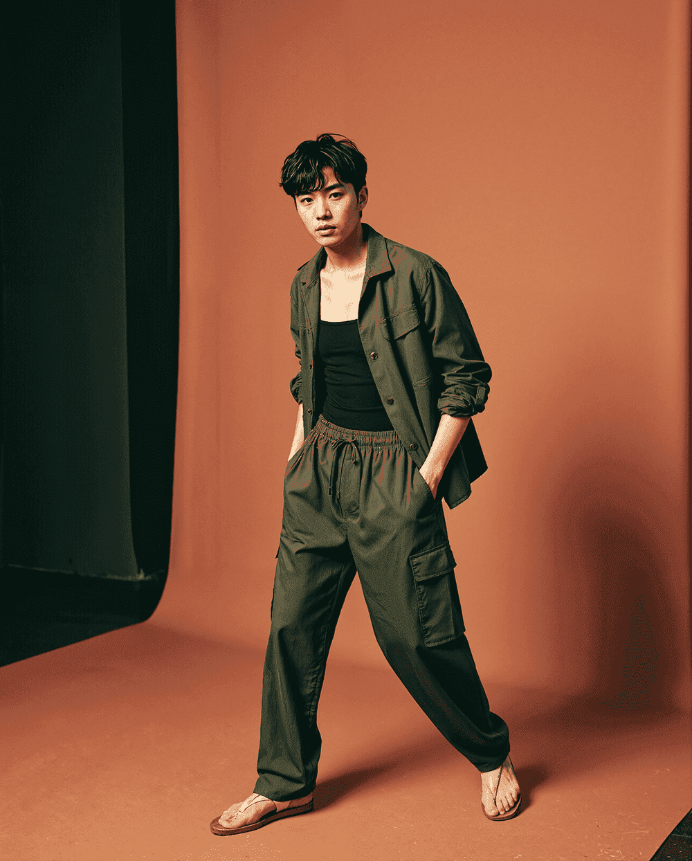 Young man in olive green cargo pants with matching overshirt and black tank top, casual streetwear fashion look styled with sandals against an orange studio background.