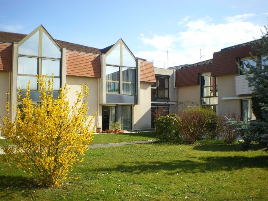 Modern residential complex with distinctive triangular glass facades, terracotta roofs, and a landscaped lawn with flowering bushes in the foreground.