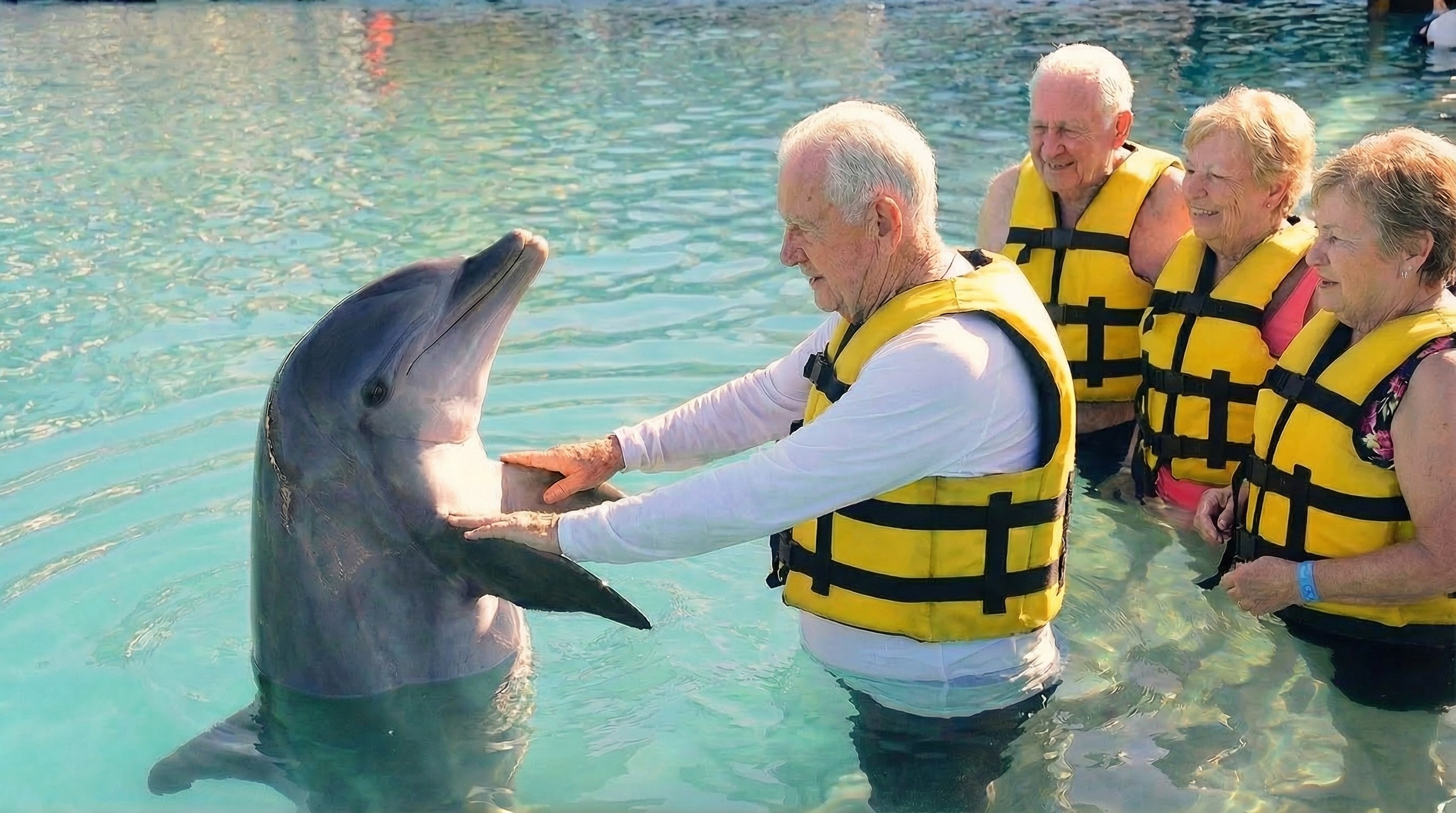 Elderly man in a yellow life vest petting a dolphin.