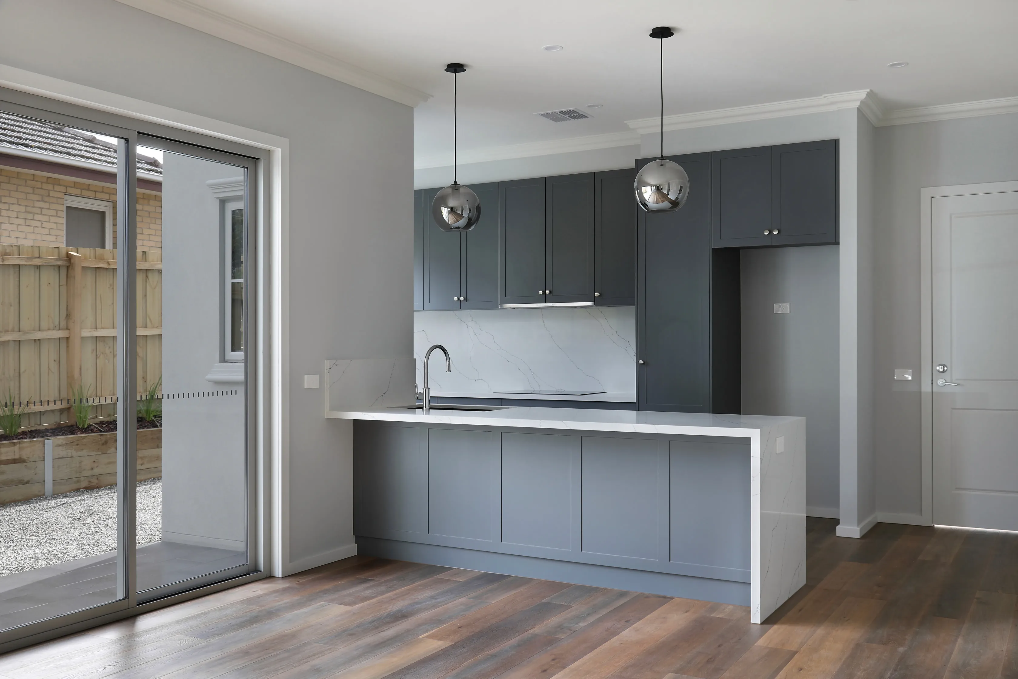 Kitchen with charcoal shaker cabinetry, a Calacatta marble splashback and white waterfall island, chrome sphere pendants and oak floors, beside a sliding door to a courtyard