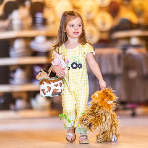 એક નાના બાળીના પીઓયલ ચેકડ કપડામાં પ્લાશ ટોય અને એક નાનું ગાયના નકશામાં બેગ પકડીને, ટોપીઓની શેલ્ફ સાથેની દુકાને ઊભું છે.