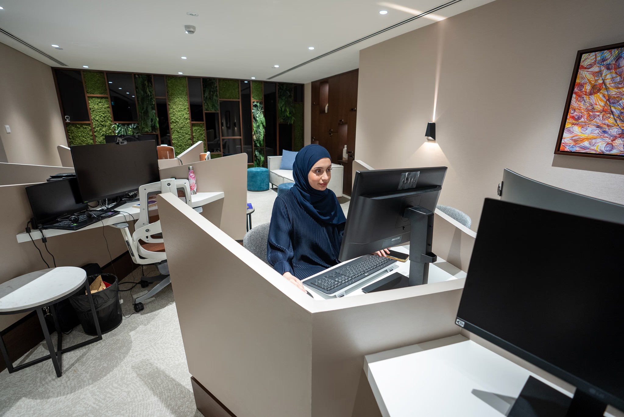 سيدة أعمال محجبة تعمل بتركيز أمام شاشة الكمبيوتر في مكتب عصري بدبي، مجهز بنباتات ولوحات فنية لإصدار رخصة تجارة الكترونية دبي