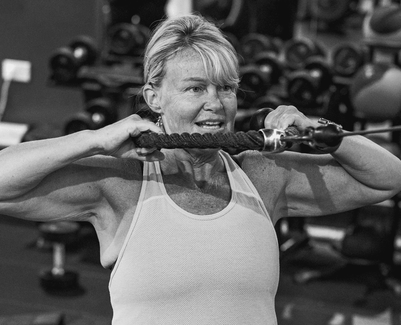 A woman is performing a cable face pull exercise in a gym with dumbbells in the background. 