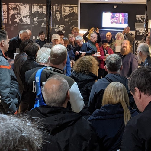 A group of people gathered in a room, some holding records, with black-and-white photos and a lit display screen in the background.