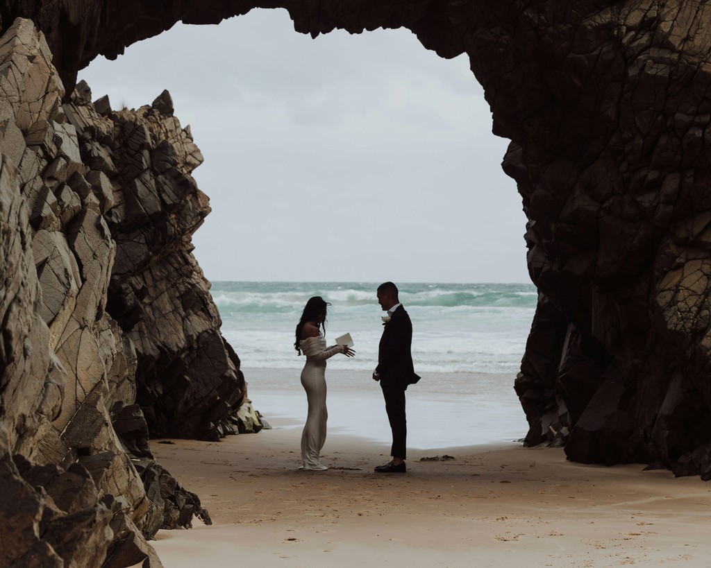 Couple exchanging vows inside a dramatic sea cave with crashing waves and a sandy beach framed by the rocky archway