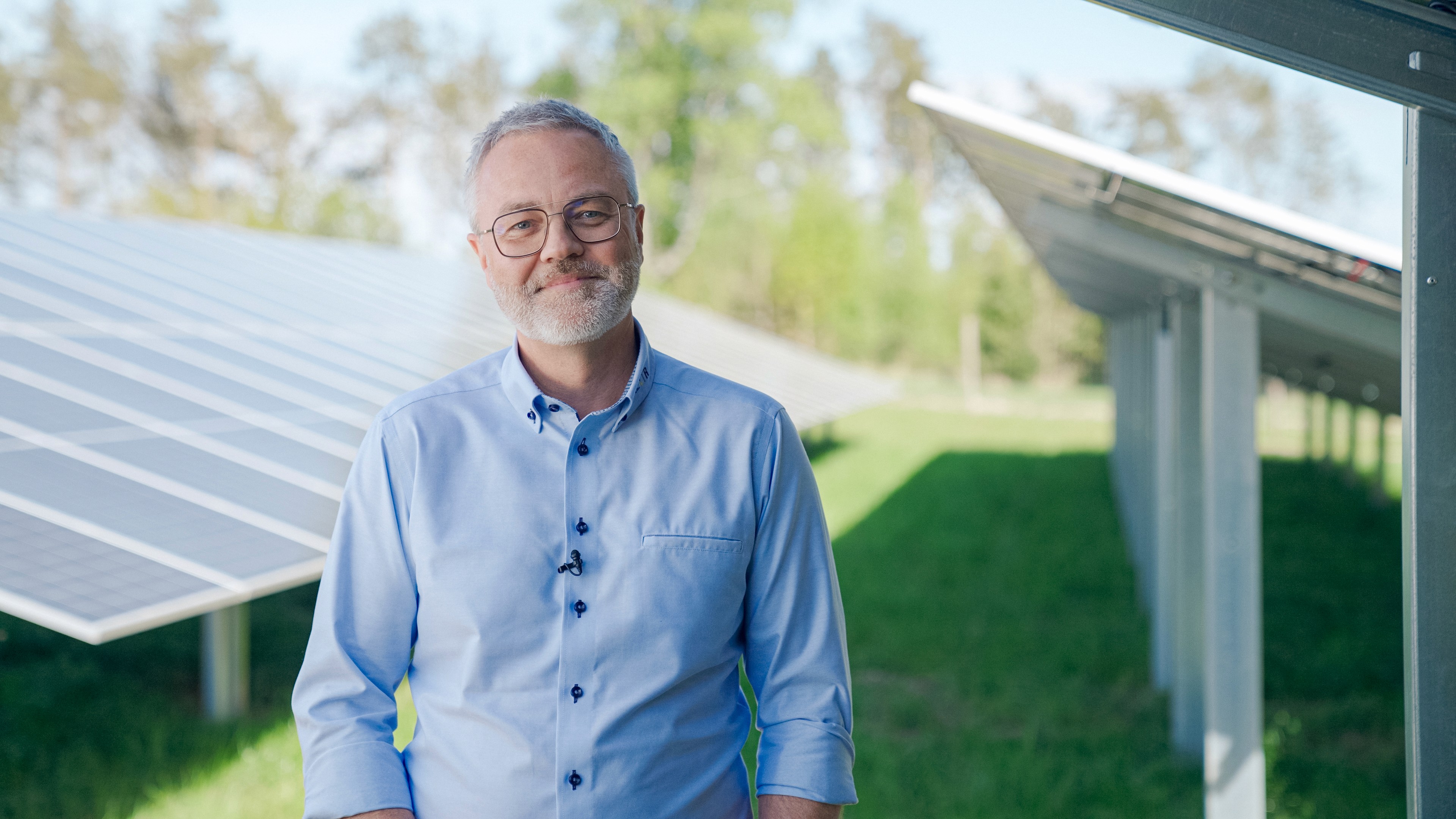 Man with white beard and glasses, wearing blue dress shirt, smiling and standing in front of a row of solar panels.