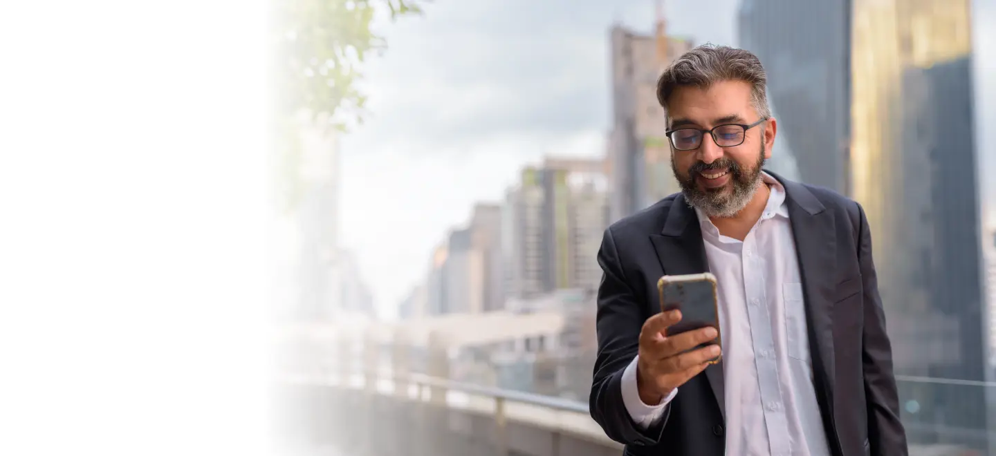 A middle-aged Indian businessman in a suit smiling while using his smartphone outdoors, representing the professional and convenient use of digital health platforms for managing chronic conditions.