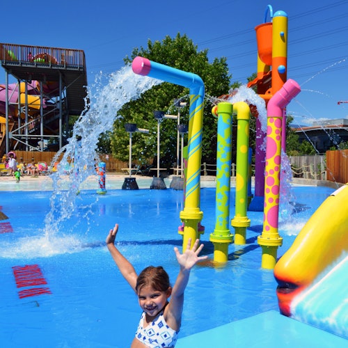 A girl smiles and raises her hands in a water park with colorful water sprays and structures in the background.
