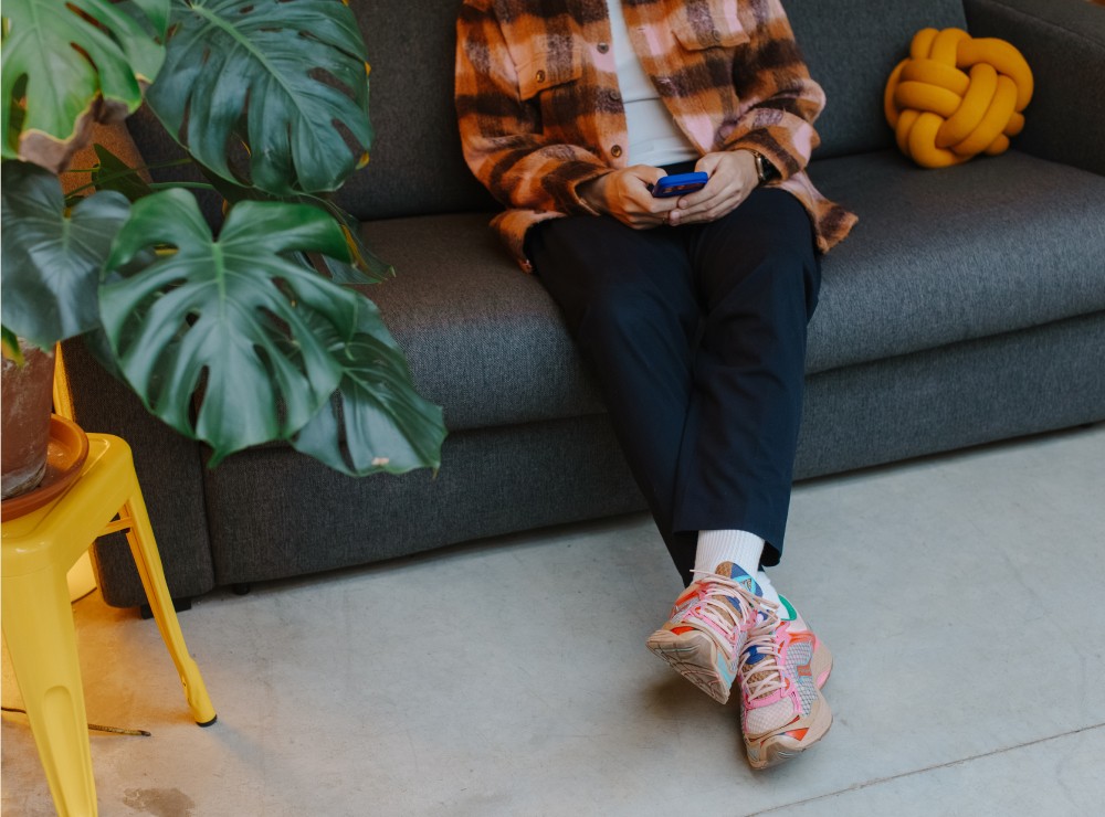 Kevin sitting on a grey sofa scrolling on their phone, with colourful sneakers and a monstera plant nearby.