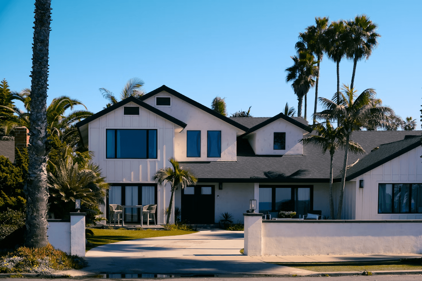 Contemporary white Spanish-style house with dark wood accents, palm trees, driveway, and lush green lawn under blue sky.
