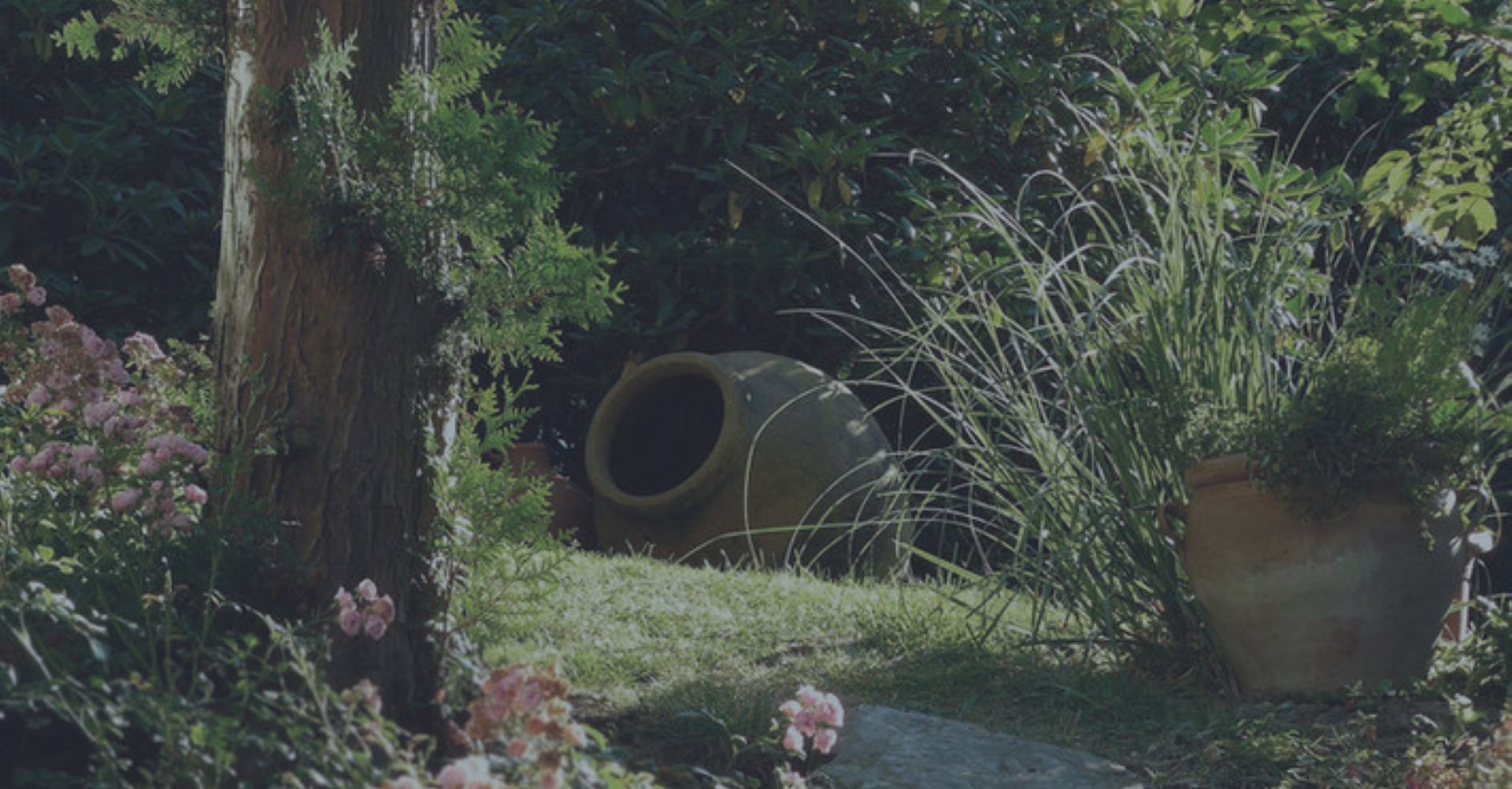 Stimmungsvolle Gartengestaltung mit einer großen Ton-Amphore, hohen Ziergräsern und blühenden Sträuchern im sanften Sonnenlicht – Gartengestaltung Klingenstein.