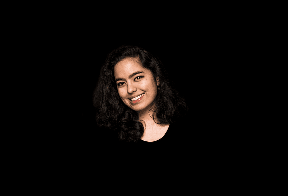 A woman with dark, curly hair is smiling at the camera against a solid black background. She is wearing a black top and looking directly at the camera.