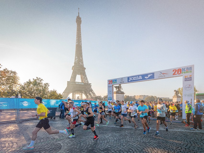 Des coureurs prenant le départ de la course des 20 km de Paris, avec la Tour Eiffel en arrière-plan et un arc de départ marqué.