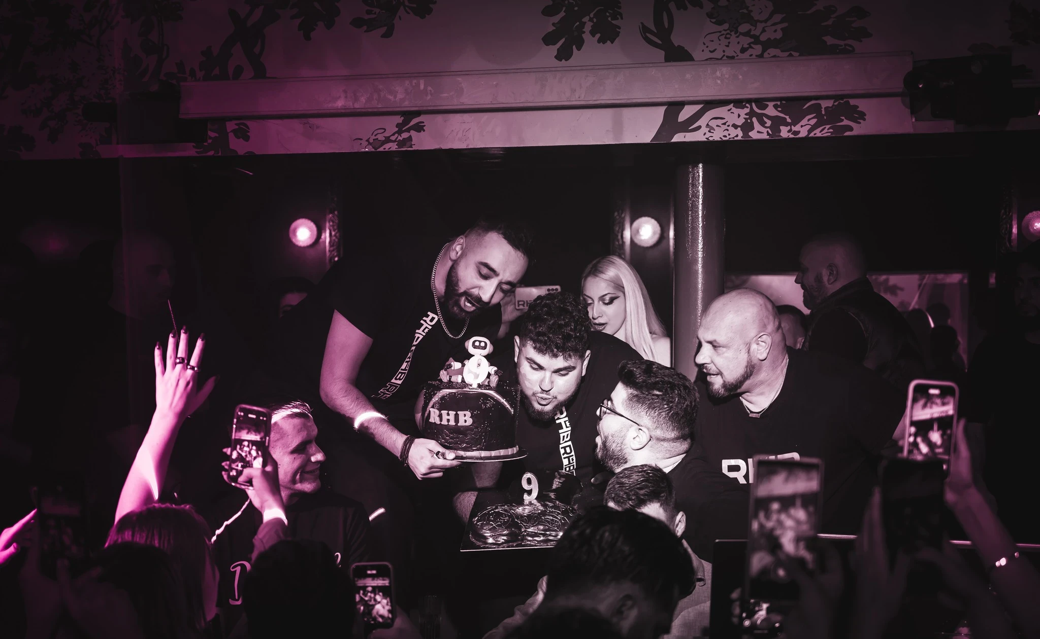 A group of people at the RHB party celebrating around a two-tiered chocolate cake. The cake features an "RHB" logo and the number 9, and other partygoers are filming with their phones.