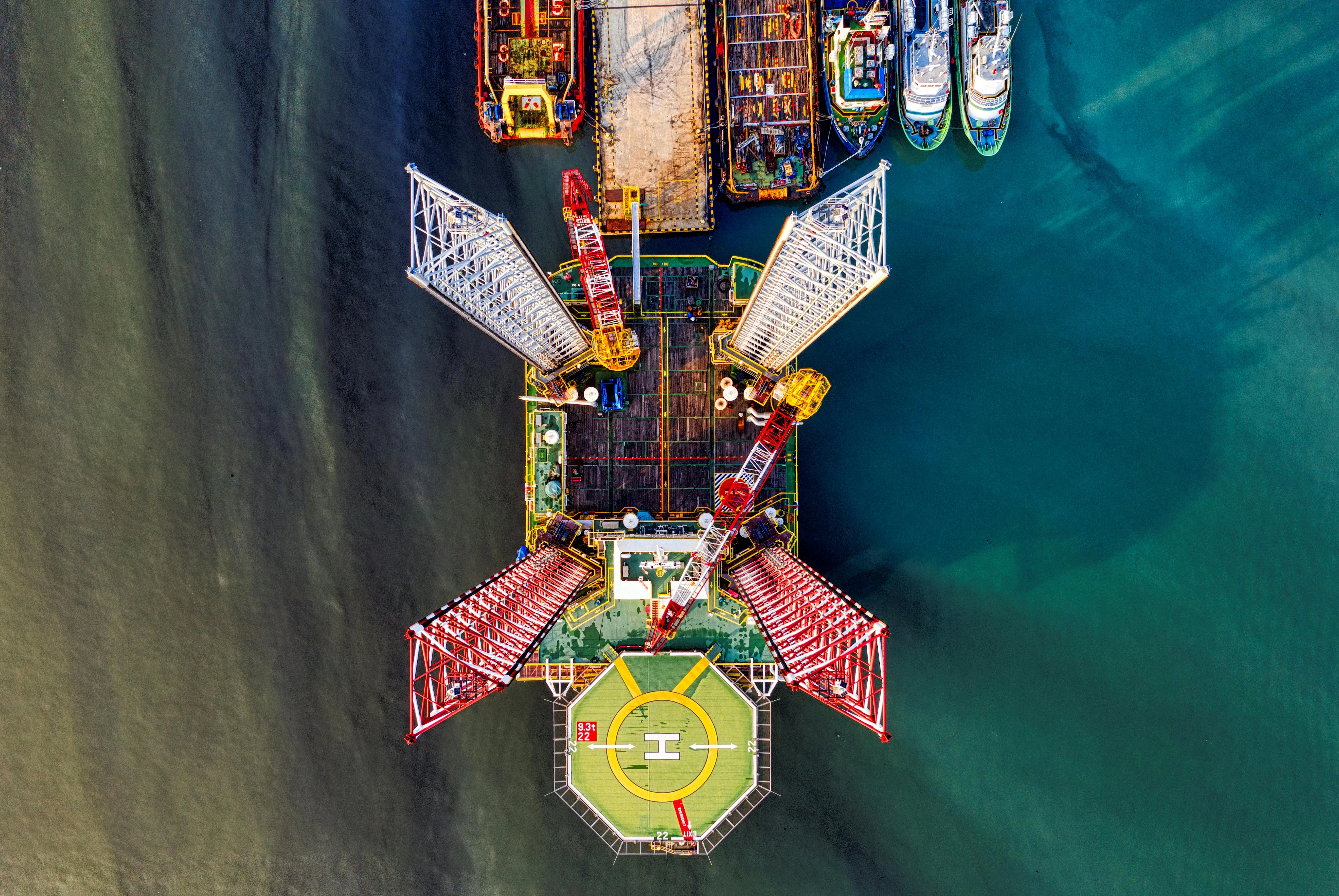 Aerial bird's-eye view of a jack-up rig with a yellow helideck docked at a port next to support vessels.