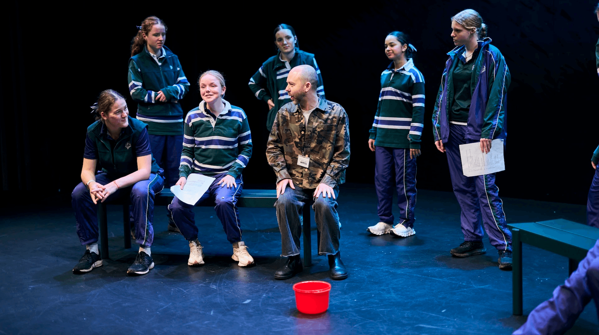 A group of teenage performers in matching green-striped school sports uniforms stand and sit around a man in a camouflage shirt, acting in a scene on a dark theatre stage. One student sits next to him holding a script, while others watch intently. A small red bucket sits on the floor in front of them.