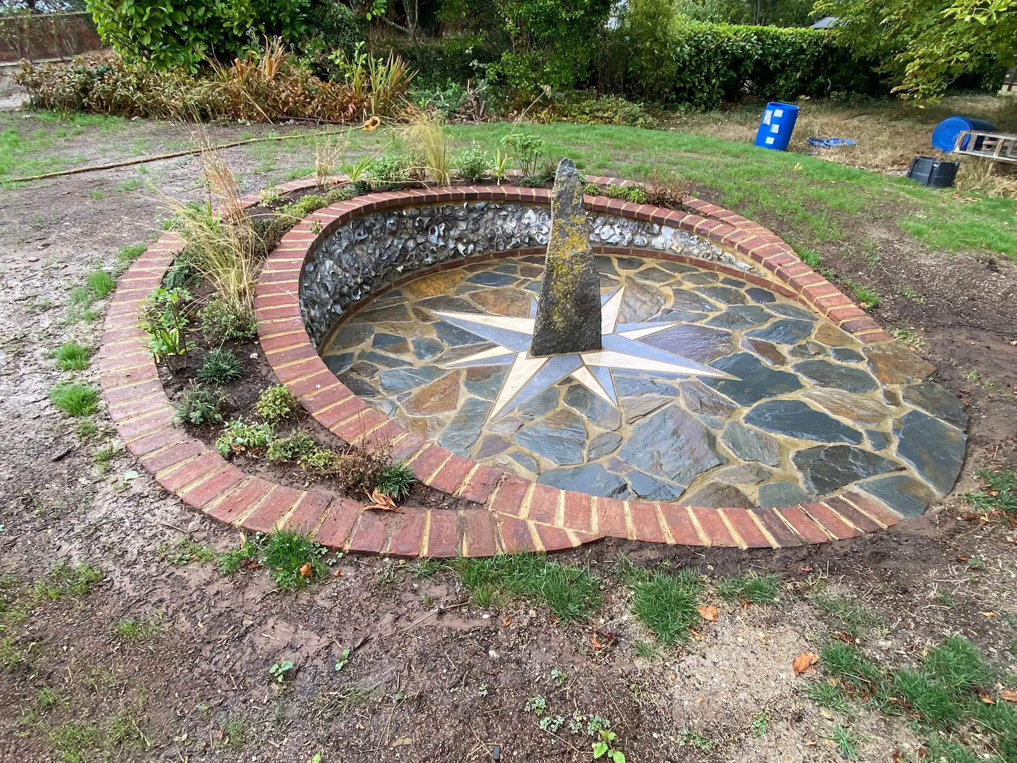 A circular stone feature with water flowing, surrounded by grass and shrubs in a garden setting.