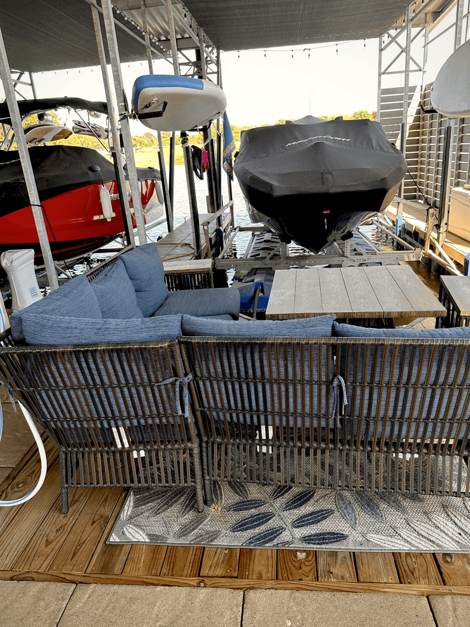 A docked area with two covered boats under a sheltered awning, featuring a cozy seating arrangement with blue cushions and a wooden table on a decorative rug, set atop a wooden deck.