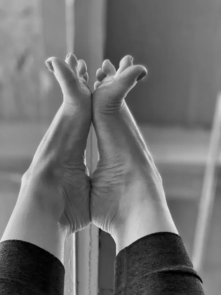 Black and white image of a person practicing yoga with their feet pressed together, raised into the air against a blurred indoor background, emphasizing flexibility and tranquility.
