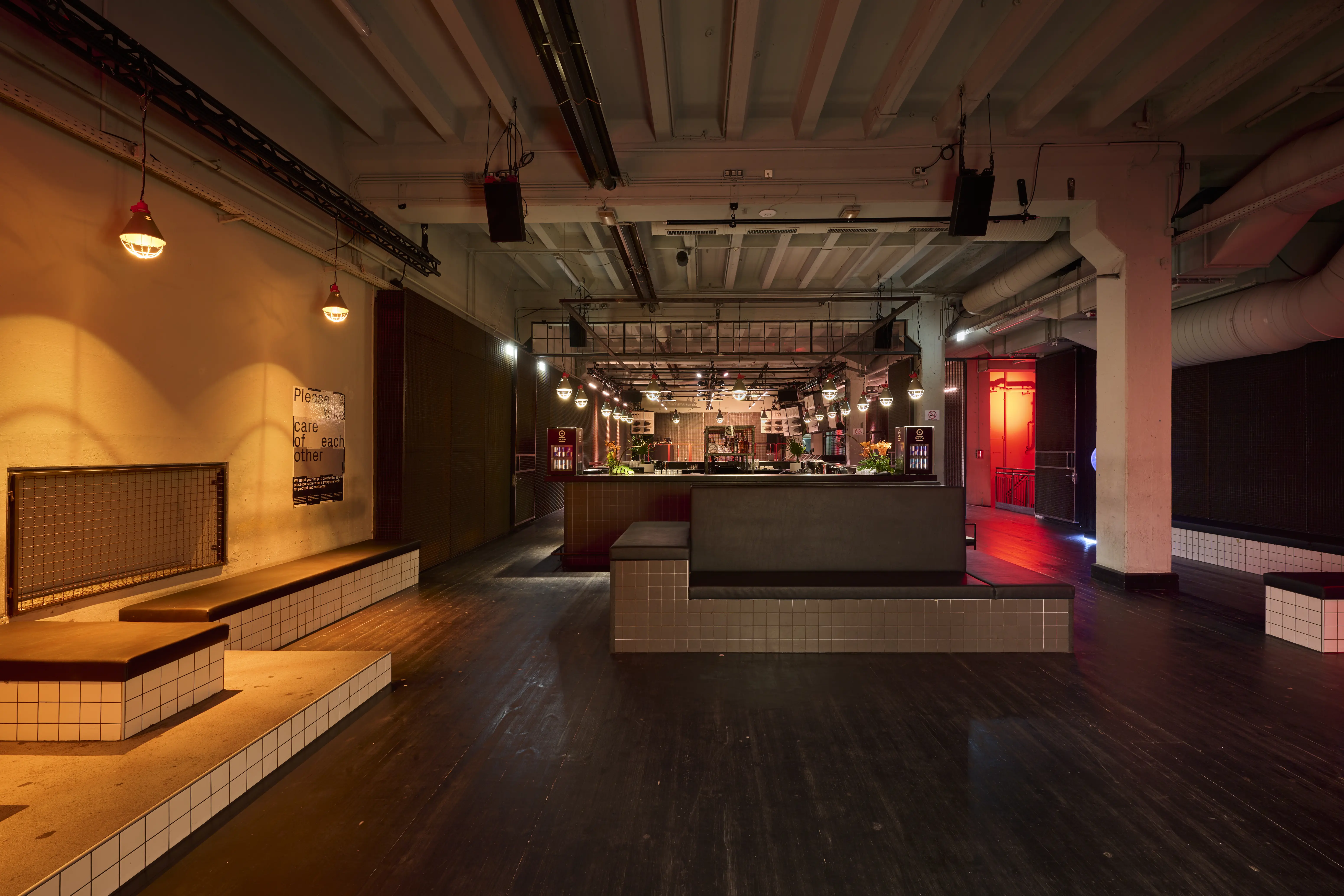 Foyer level at TRESOR GLOBUS with white-tiled seating platforms with dark cushions, warm wall lights, a central tiled bar block and a red-lit door in the background.