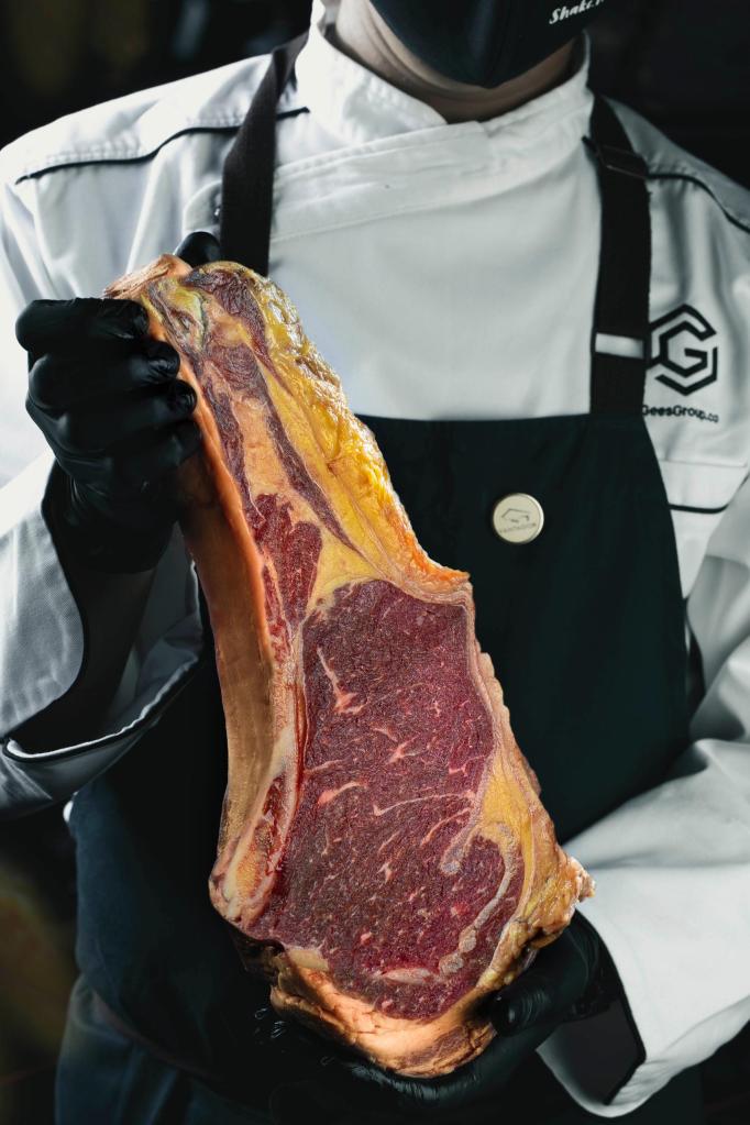 A chef in a white uniform and black gloves holding a large cut of aged beef ribeye, showcasing its marbling and color.